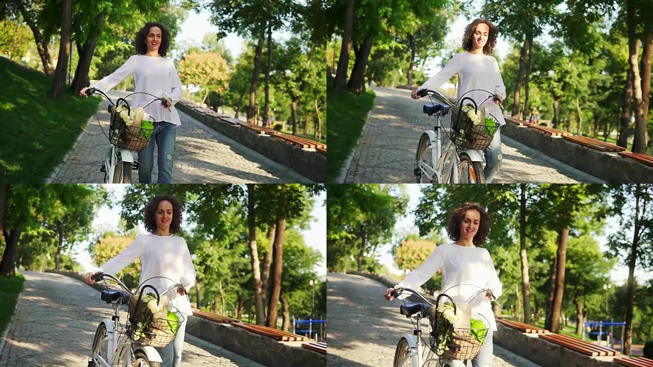 清晨，穿着白色t恤和蓝色牛仔裤的微笑迷人的女人拿着城市自行车的车把，篮子里放着鲜花。快乐的女人享受她
