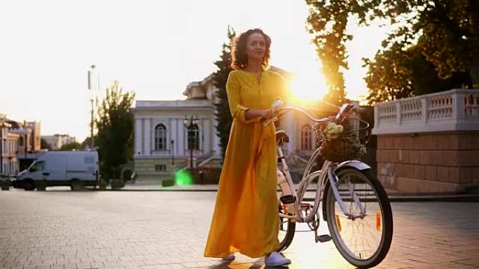 穿着黄色长裙微笑的黑发女人在黎明时拿着城市自行车的车把，篮子里放着鲜花。镜头耀斑，美丽的城市景观