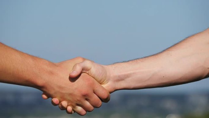 两个无法辨认的肌肉发达的白人在蓝天背景上友好握手。室外摇晃男性手臂。两个强壮的男人在外面坚定地握手。