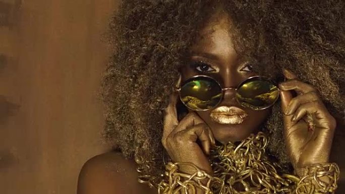 非裔美国女模特在巨大的太阳镜与明亮的闪闪发光的化妆，有光泽的金色发型和大红色的嘴唇摆姿势在黑色的工作