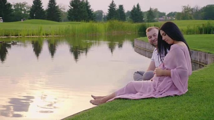 和丈夫一起穿着粉红色连衣裙的孕妇。他们坐在湖边的草地上