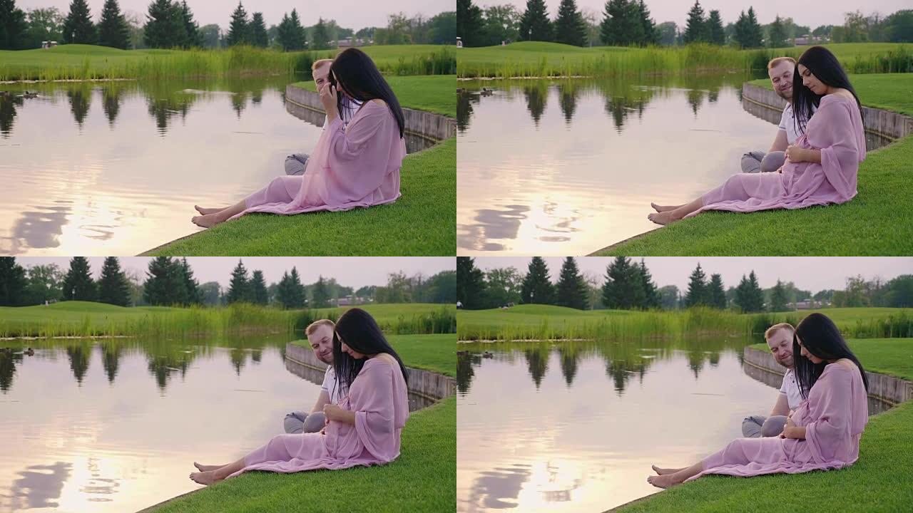 和丈夫一起穿着粉红色连衣裙的孕妇。他们坐在湖边的草地上