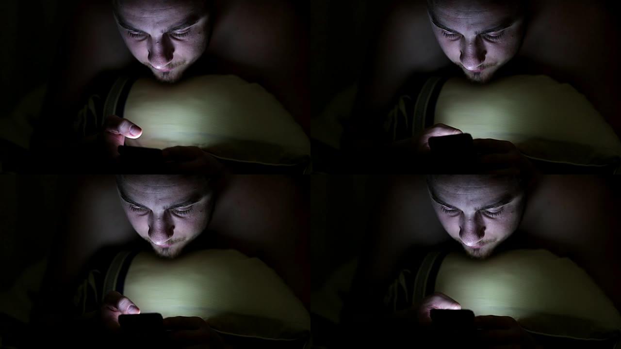 年轻人晚上在毯子下的床上使用智能手机