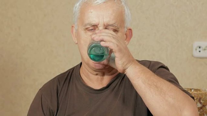 生病的超重老人在沙发上的房子里吃药，在杯子里喝水。他制造了几种光滑的液体。老年人的健康观念