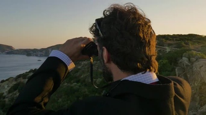 穿着优雅西装的男人，用公文包在岛上的悬崖顶上透过双筒望远镜看