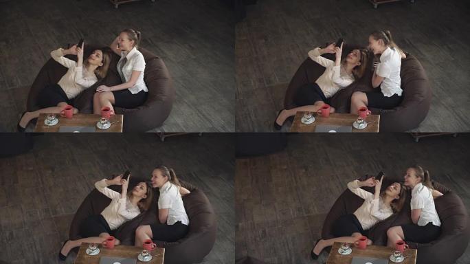 两个激动人心的金发女郎躺在沙发上，在手机上查看照片，讨论快速制作的自拍照片，大声笑