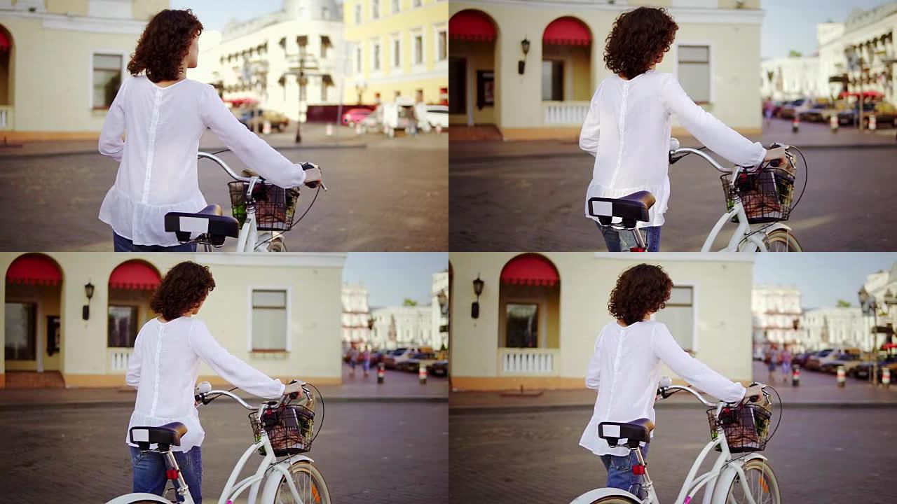 一个穿着白色衬衫和蓝色牛仔裤的女人在城市街道上行走的背景图，她拿着自行车的车把，篮子和鲜花。清晨美丽