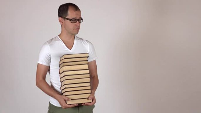 戴着黑框眼镜的书呆子男人在灰色背景下背着一大堆书，与世隔绝。全高清镜头