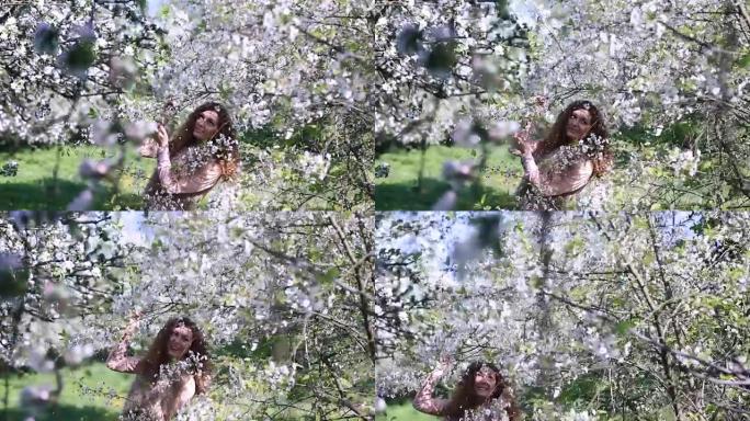 年轻的女孩在春天的果园里摇晃着盛开的樱桃枝，当花瓣像雪花一样落下时大笑