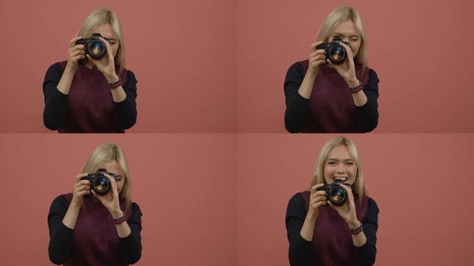 迷人的金发女人用相机拍照，在粉色背景下大笑