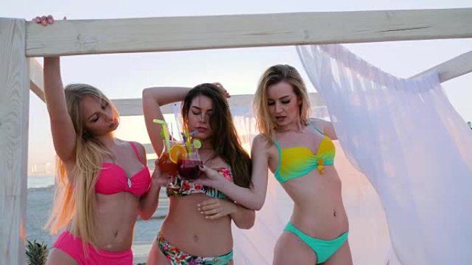 女友在背光的海滩上跳舞，诱人的女人喝酒精鸡尾酒，性感的泳衣