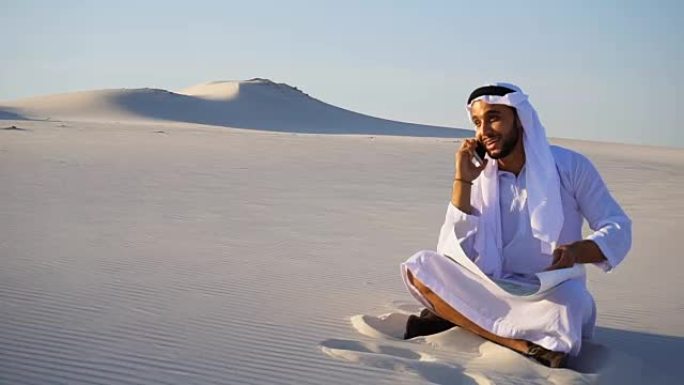 在炎热的日子里，一个自信的阿拉伯阿拉伯建筑工人坐在沙地上用手机与客户交谈