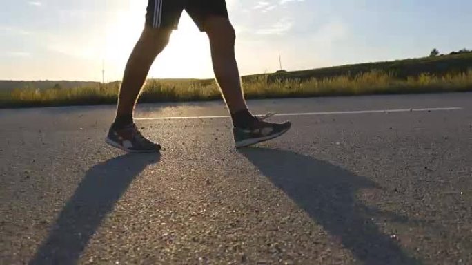 男性的脚在日落时在乡村道路上行走，靠近。年轻人正走在一条空旷的乡间小路上。穿过阳光照射的人。小男孩的