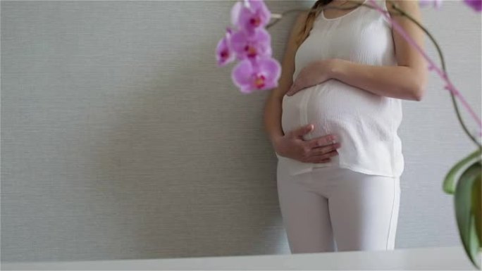 美丽的年轻孕妇正站在灰色的墙壁上抚摸着一个大肚子。