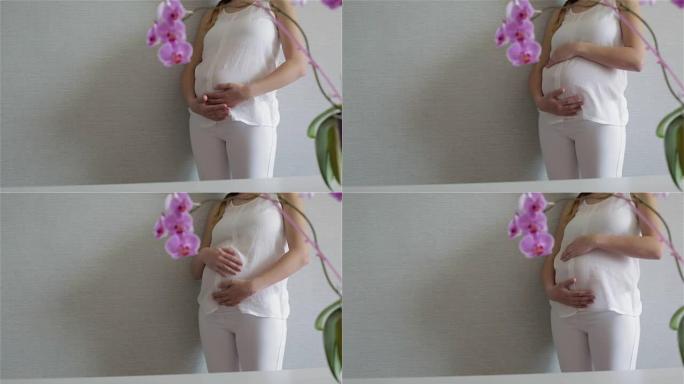 美丽的年轻孕妇正站在灰色的墙壁上抚摸着一个大肚子。