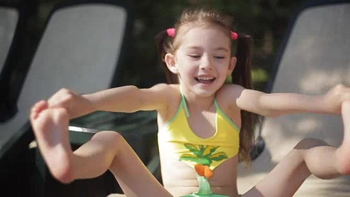 一个穿着泳衣的小女孩坐在游泳池边的躺椅上笑着。