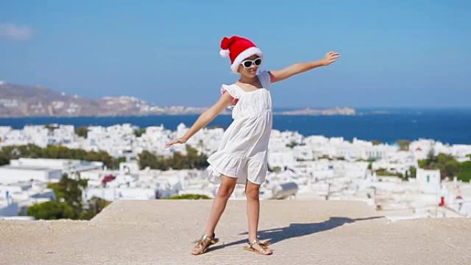 米科诺斯户外背景的红色圣诞老人帽子里有趣的小女孩。希腊圣诞节假期，孩子在典型的希腊传统村庄的街道上，