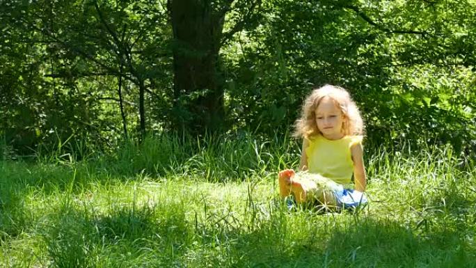 美丽的小女孩的户外肖像，金色的短卷发坐在绿色的草地上，捡起并扔掉。
