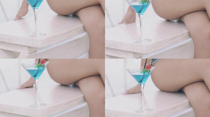 靠近裸露女人的腿和手的蓝色鸡尾酒杯。慢慢地