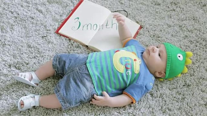 戴着绿龙帽子的三个月婴儿躺在他背上的灰色地毯上。