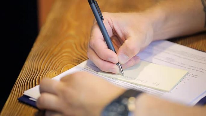 男子坐在木桌旁，用中性笔在贴纸上写短信