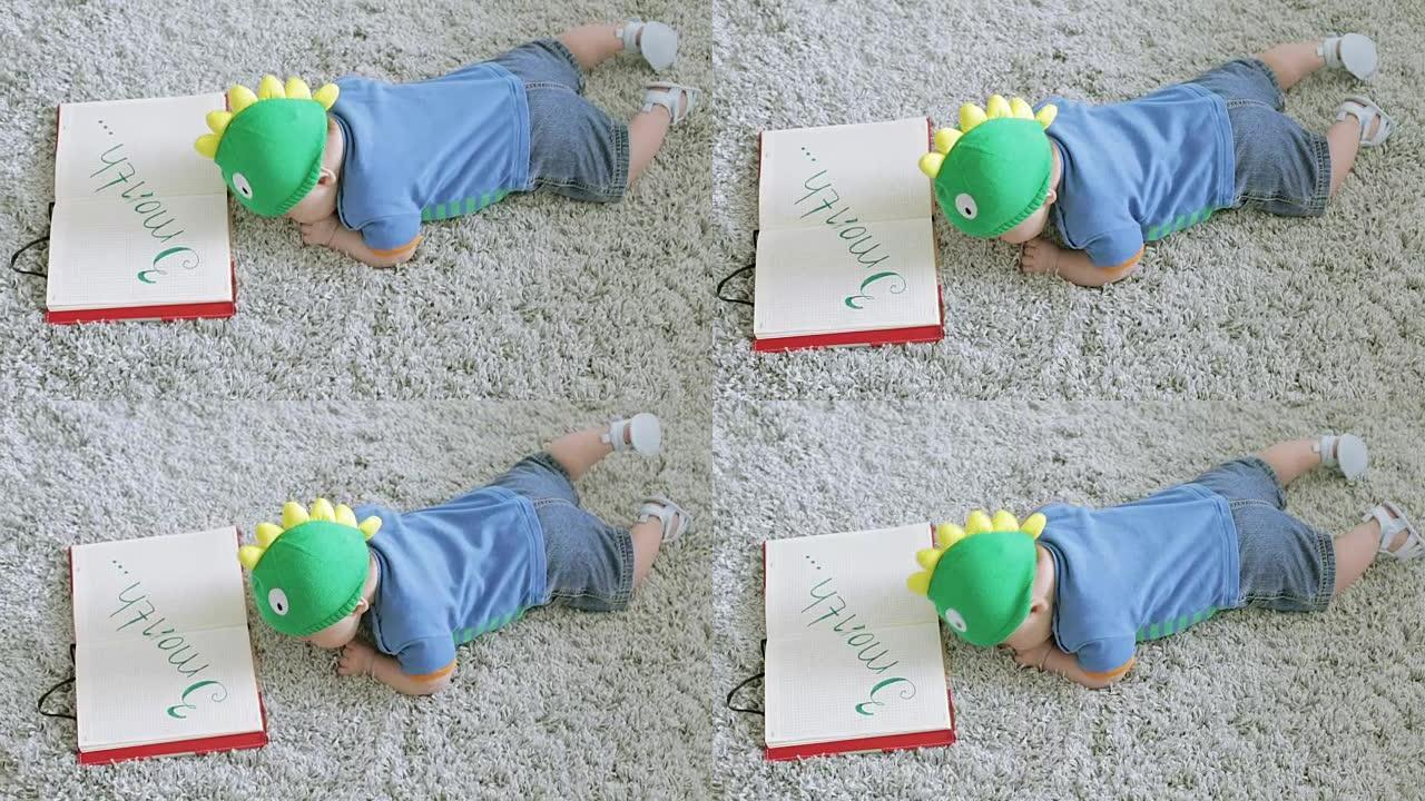 戴着绿龙帽子的三个月婴儿看着躺在地上的笔记本。
