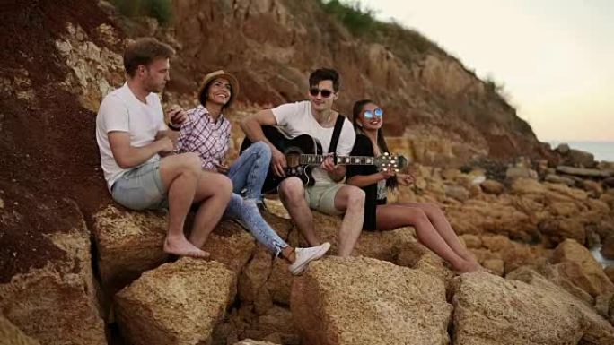 一群年轻的时髦朋友坐在海边的岩石上，弹吉他，唱歌和跳舞。慢动作镜头
