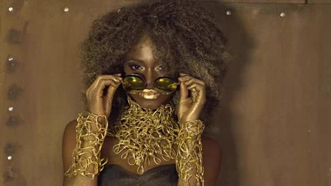 魔幻超现实的黄金非洲裔美国女模特在巨大的太阳镜与明亮的闪闪发光的化妆，有光泽的金色发型和大红色的嘴唇