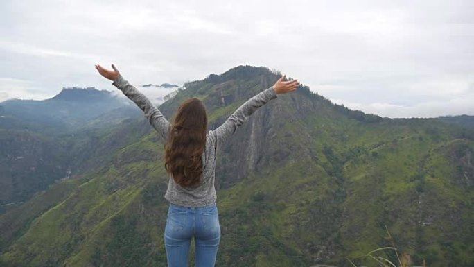年轻女孩伸手爬山，举起双手。女游客站在美丽的峡谷边缘，胜利地伸出双臂。慢动作后视图