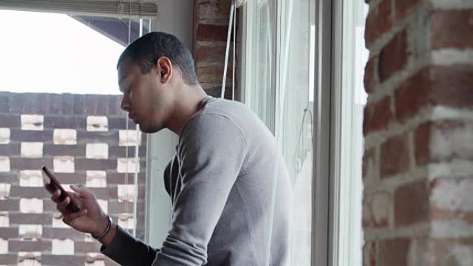 年轻的黑人男子使用智能手机放松身心，并在现代工业房屋的室外窗户室内看。4k手持特写视频