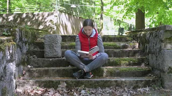 穿着红色夹克和条纹毛衣的女孩，在古老台阶上的美丽位置读一本大书。慢动作。