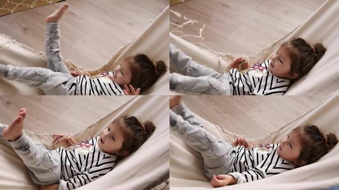 快乐的小女孩在室内玩得开心，躺在吊床上，挥舞着双腿。微笑的孩子