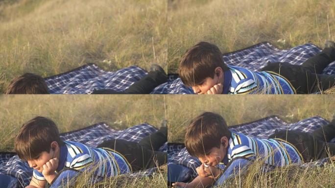 小男孩看智能手机男孩晚上躺在草地上的地毯上，挥动双腿