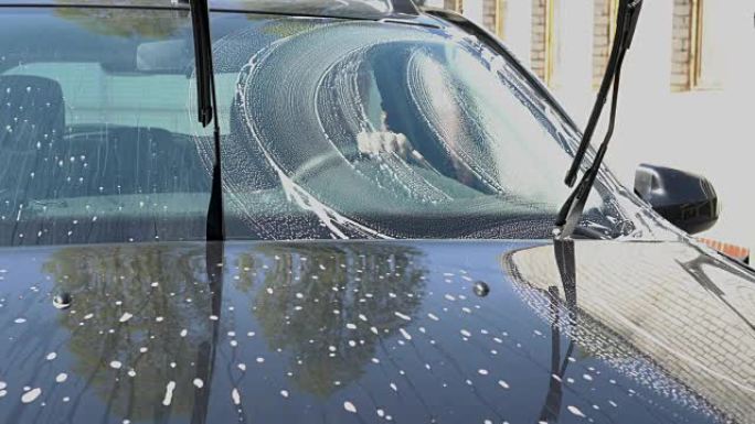 女孩在晴天用海绵洗黑色汽车。她正在洗挡风玻璃上的肥皂泡沫。方向盘后面的车内坐着一个男人，透过泡沫看着