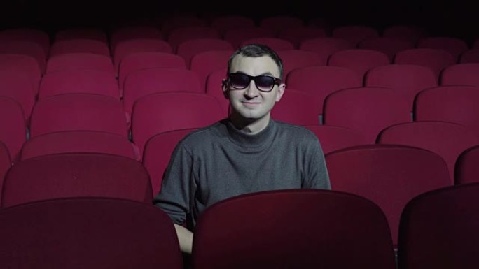 穿着3d眼镜的单身男子坐在黑暗电影院剧院舒适的红色椅子上，看着相机，微笑着