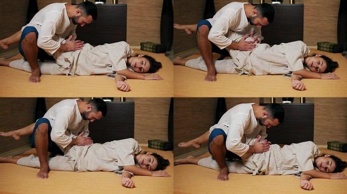 泰国男性按摩师正在治疗年轻女子的臀部和坐骨神经，握住她的腿并伸展它们。年轻迷人女性的泰式按摩课程