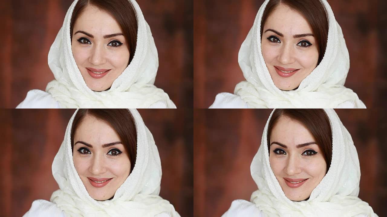 一位年轻而神秘的阿拉伯妇女的特写肖像穿着白色头巾，被称为头巾，微笑着看着相机。深棕色背景上美丽的穆斯