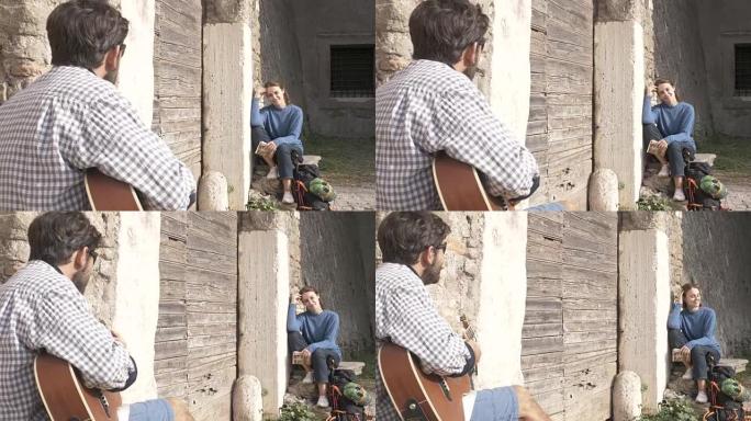快乐的年轻夫妇背包客游客弹吉他唱歌坐在罗马帕尔科德格里阿奎多蒂公园遗址中世纪建筑的木制门口附近，日出