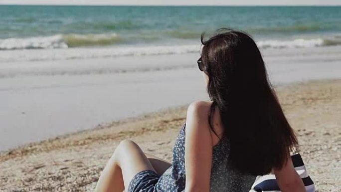 穿着蓝色牛仔连身裤的美丽年轻黑发女人坐在海岸线的沙滩上，看着海洋地平线。暑假期间海滩上的年轻女子。夏