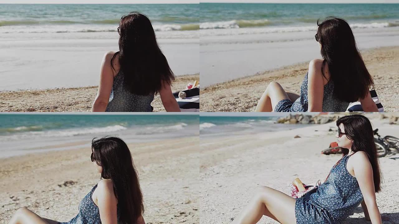 穿着蓝色牛仔连身裤的美丽年轻黑发女人坐在海岸线的沙滩上，看着海洋地平线。暑假期间海滩上的年轻女子。夏
