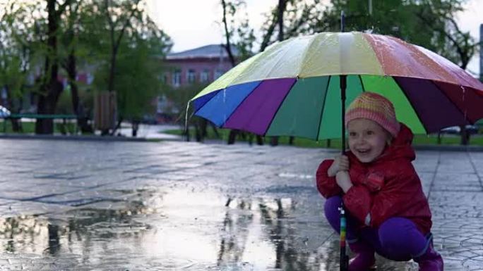 带着五颜六色伞的可爱的小女孩在日落时在公园的水坑里跳跃。