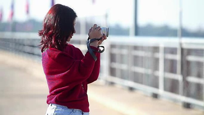 穿着红色连衣裙的年轻亚洲女孩在大坝上拿着相机拍照
