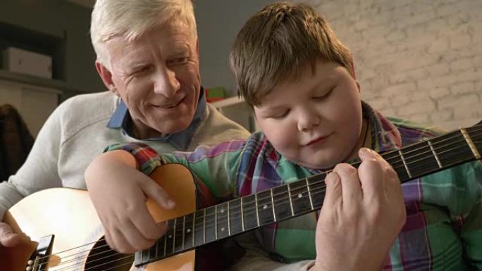 祖父正在教他的孙子如何弹吉他。一位老人正在教一个胖子弹吉他。家居舒适，家庭田园诗，舒适概念，世代差异