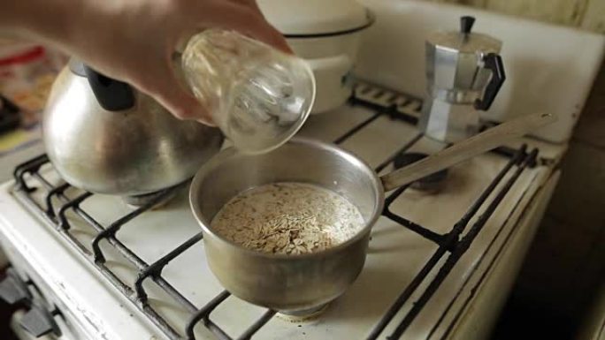 健康素食早餐，燕麦粥。一名白人妇女的手将大燕麦片从玻璃杯中倒入煤气炉上的金属锅中。强健肌肉的有机早餐