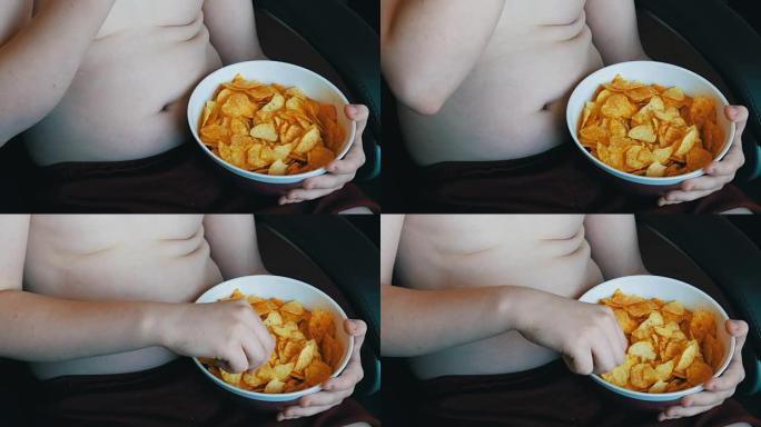 身体上有脂肪褶皱的胖少年正在用手吃薯片。不健康食品，快餐。孩子的营养问题