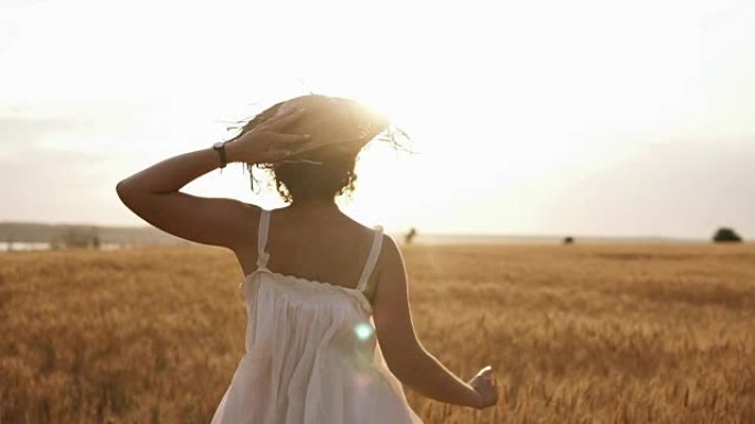 穿着草帽和夏季白色连衣裙的快乐苗条的女孩在清澈的麦田里快乐地奔跑。阳光照在背景上。背面视图