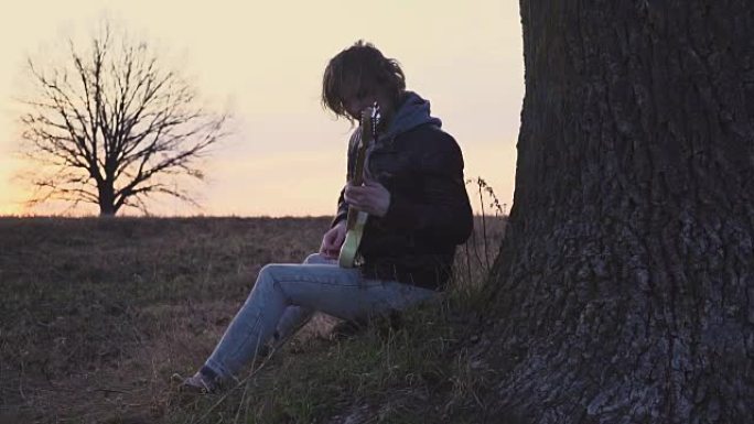 男子在日落时在树附近的田野里弹电吉他并唱抒情歌