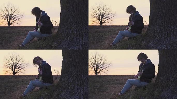 男子在日落时在树附近的田野里弹电吉他并唱抒情歌