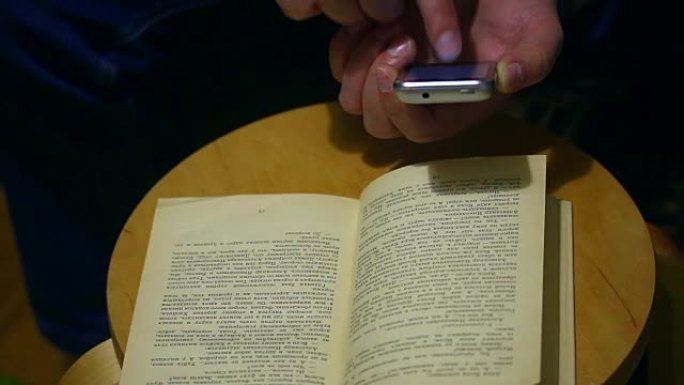 一个退休年龄的人在一本纸质书的智能手机页面上拍照。打开的书放在椅子上。这个人翻过一页。
