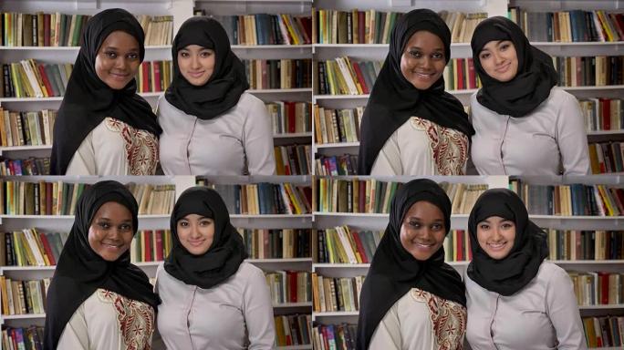 两个年轻的穆斯林妇女站在图书馆里，在镜头前微笑的肖像，伊斯兰朋友